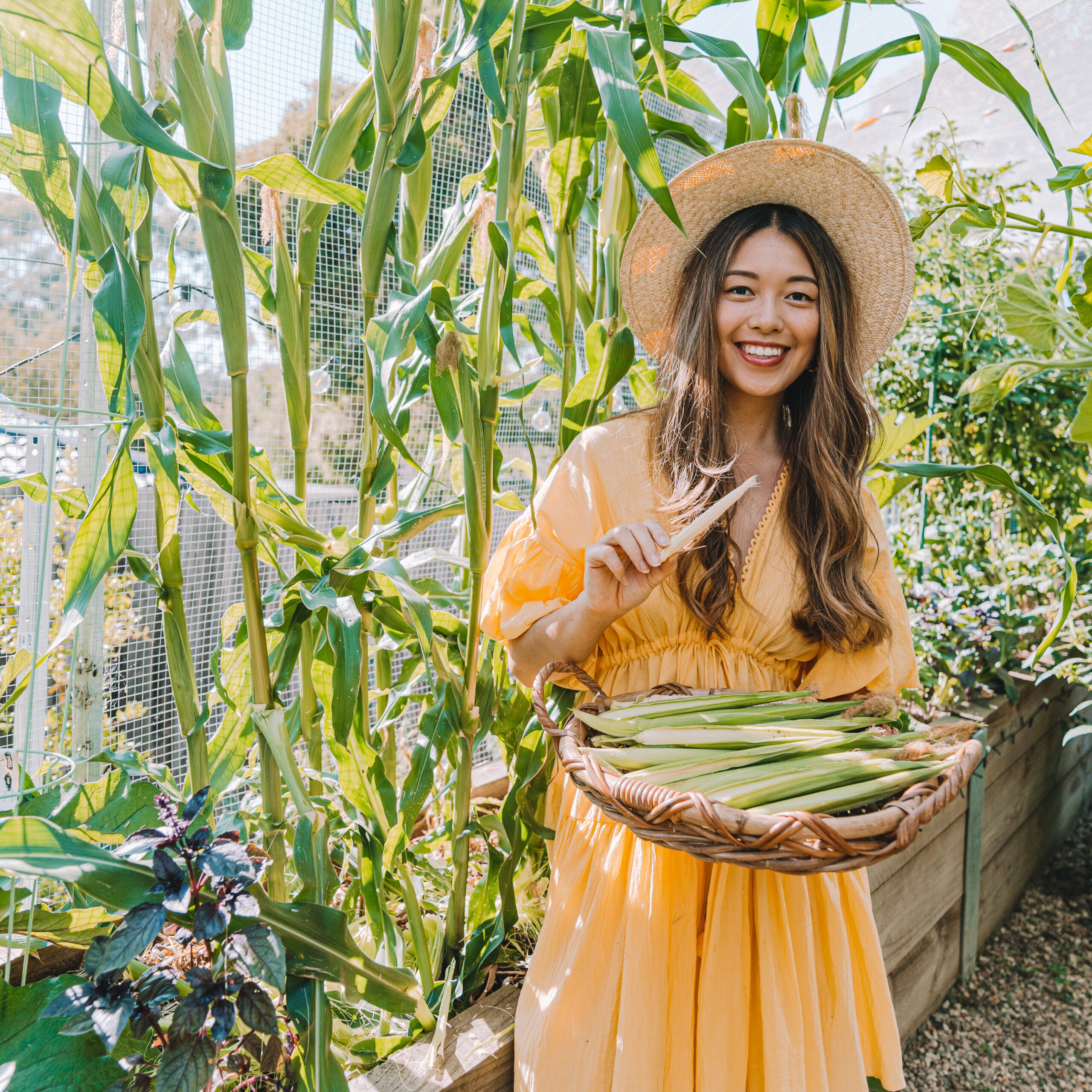 Woman in garden holding corn basket, smiling.