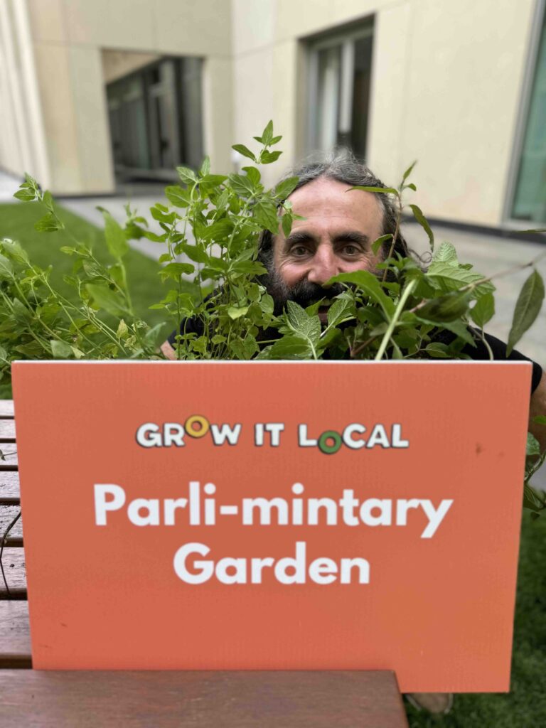 Person in garden behind 'Grow It Local' sign.