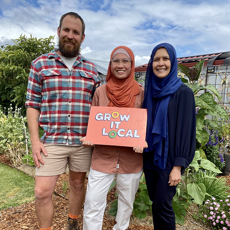 Three people holding Grow It Local sign in garden.