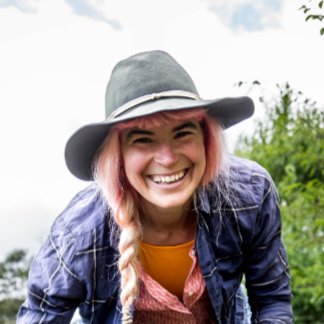 Smiling person outdoors, wearing hat and braided hair.