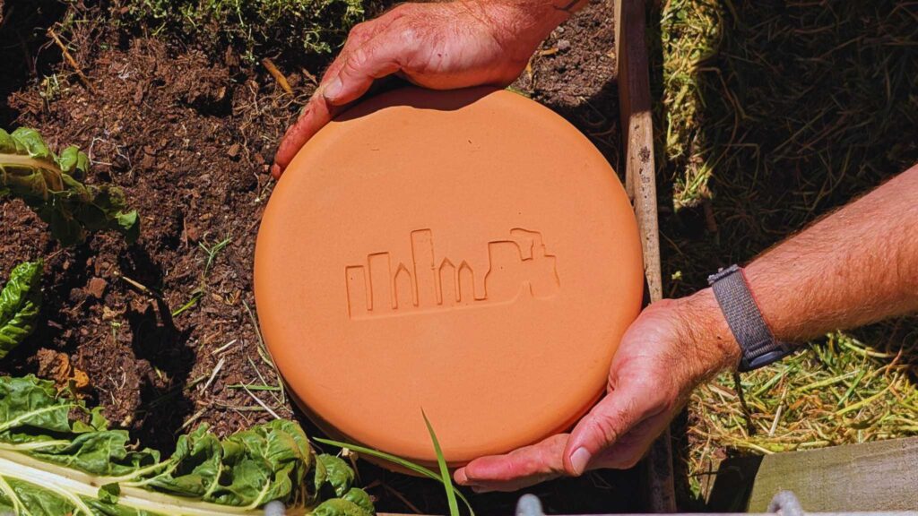 Person placing terracotta disk in garden soil.